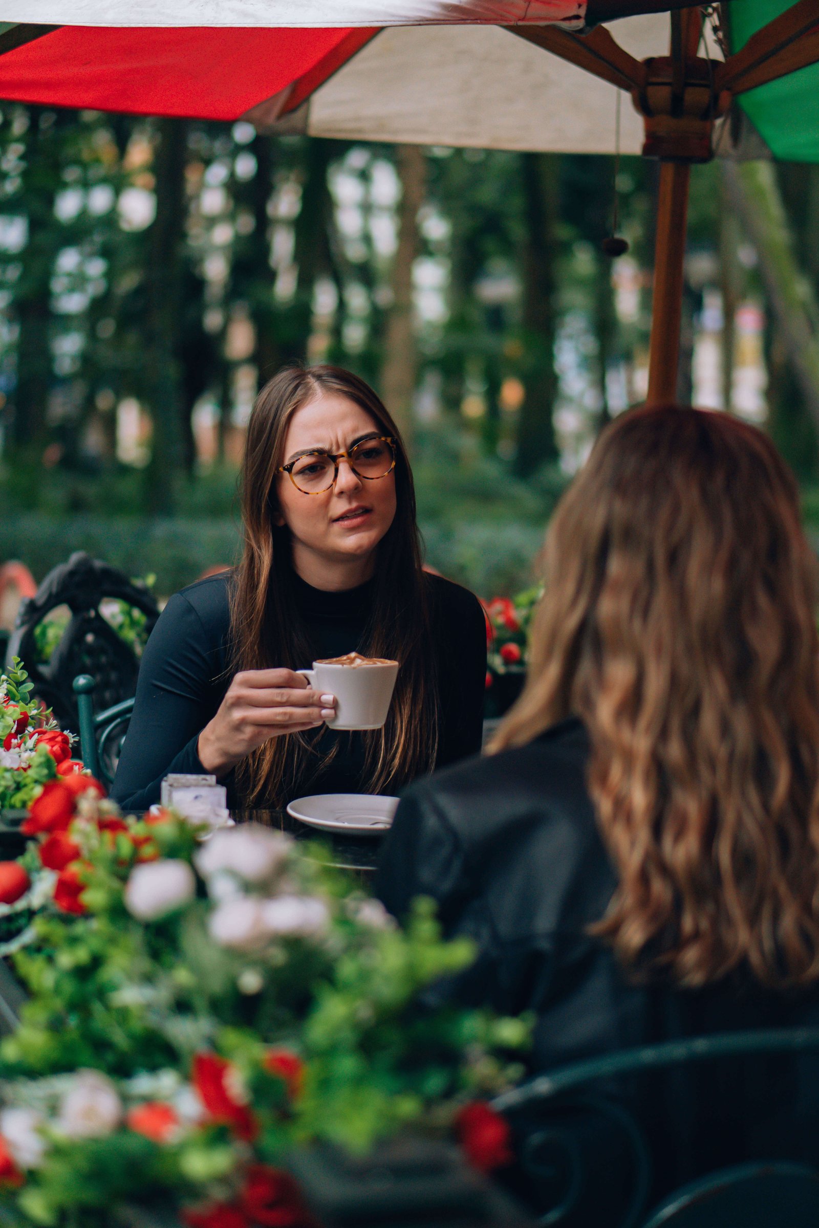 Cena de duas mulheres em mesa externa de cafeteria, uma de óculos de grau conversando entre plantas.