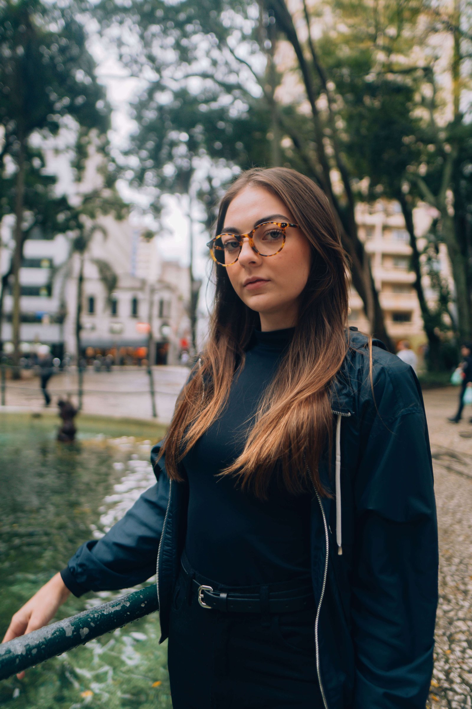 Mulher com óculos de grau e casaco escuro posando em calçada ao lado de canal e árvores.