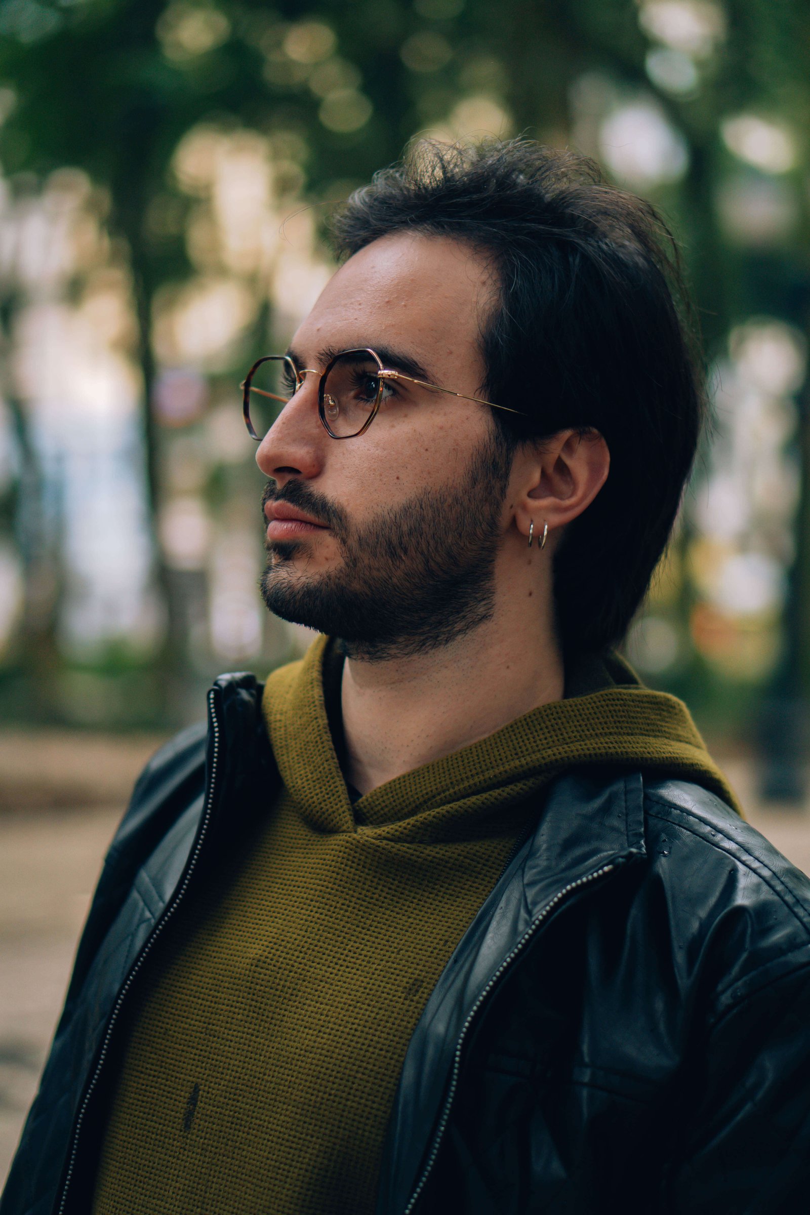 Perfil de homem com óculos de grau e barba, usando jaqueta de couro em ambiente externo.