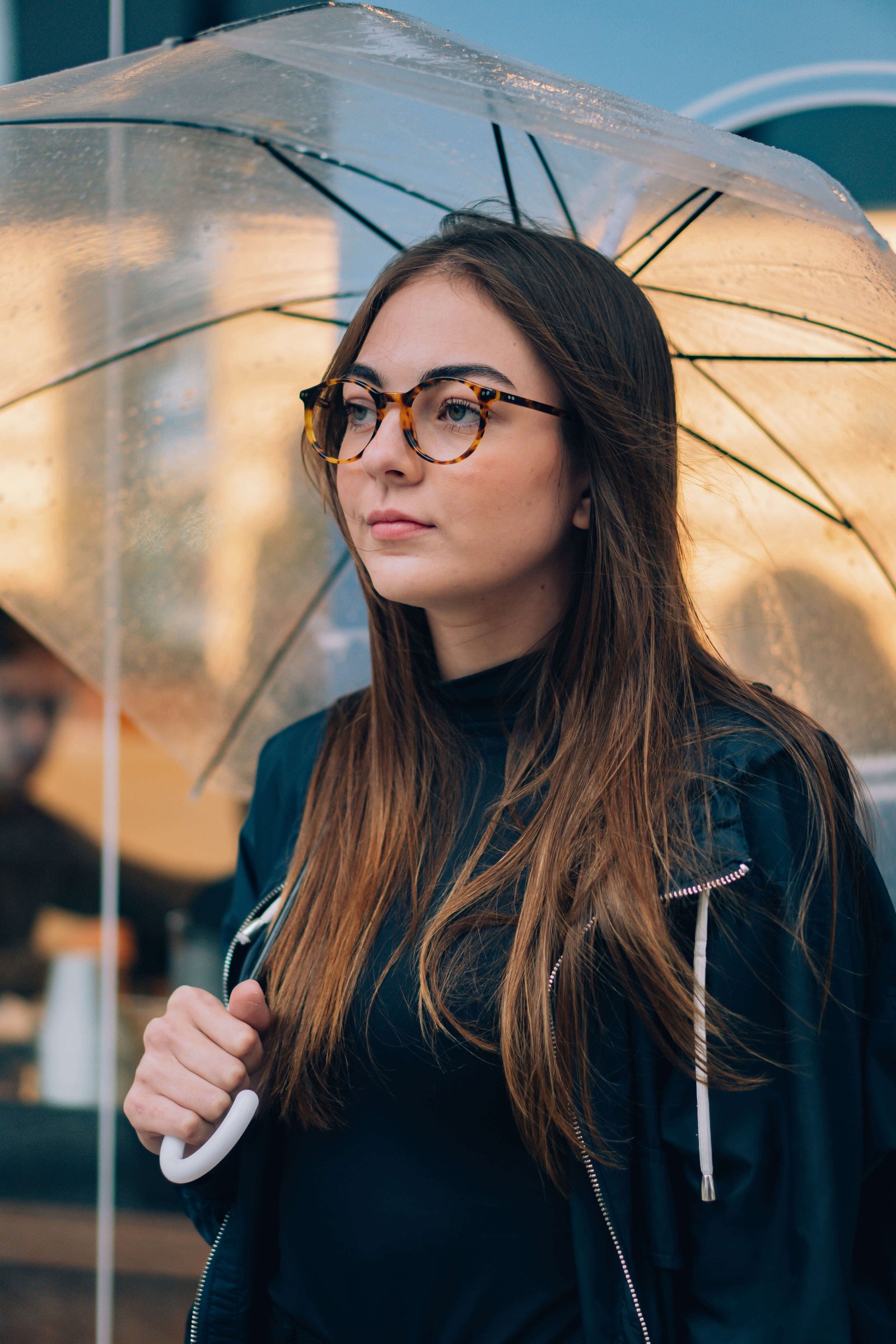 Mulher com óculos de grau segurando guarda-chuva transparente em frente a vitrine iluminada.