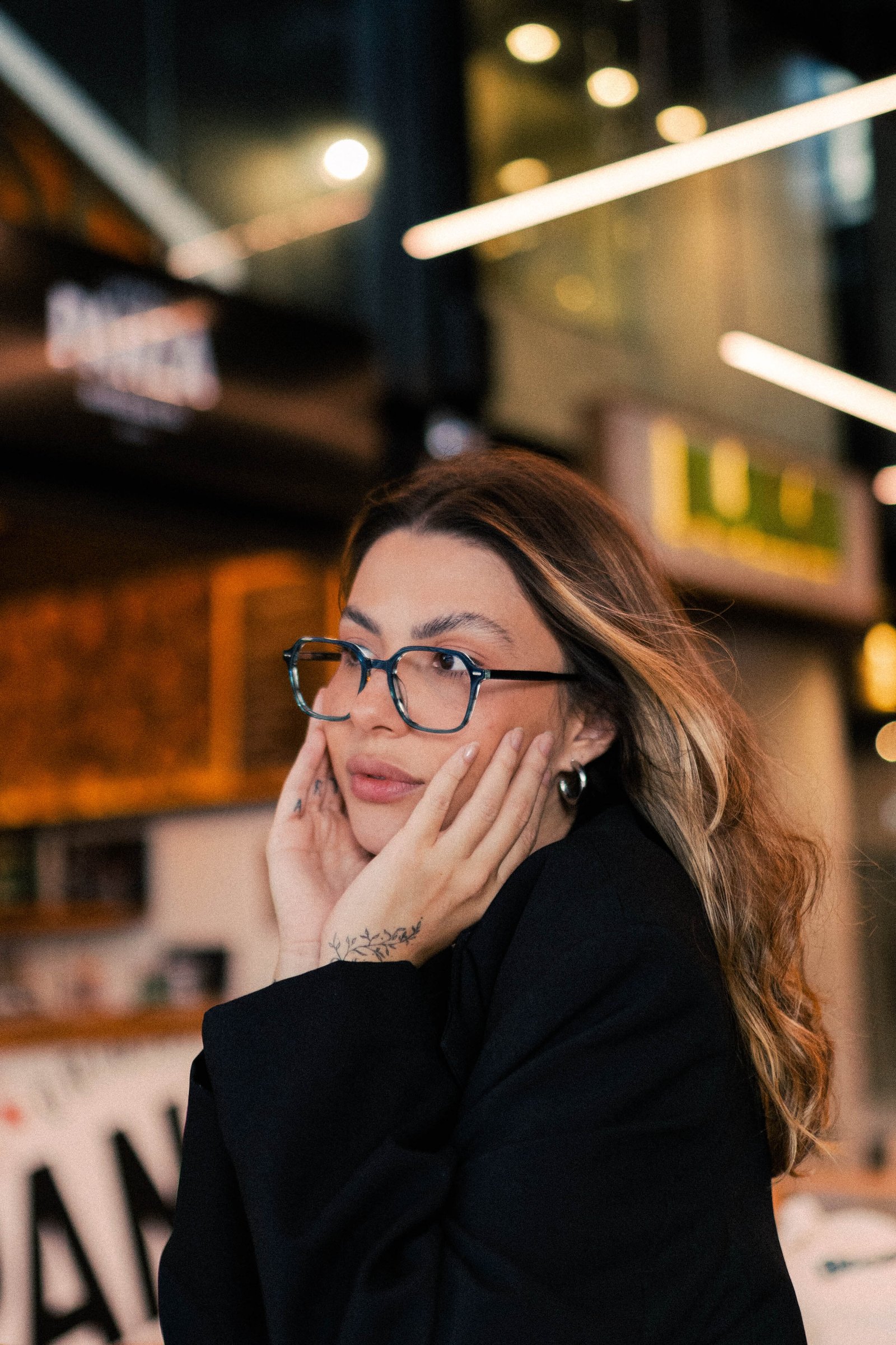 Mulher com óculos de grau apoiando o queixo na mão, sentada em mesa de café com luzes ao fundo.