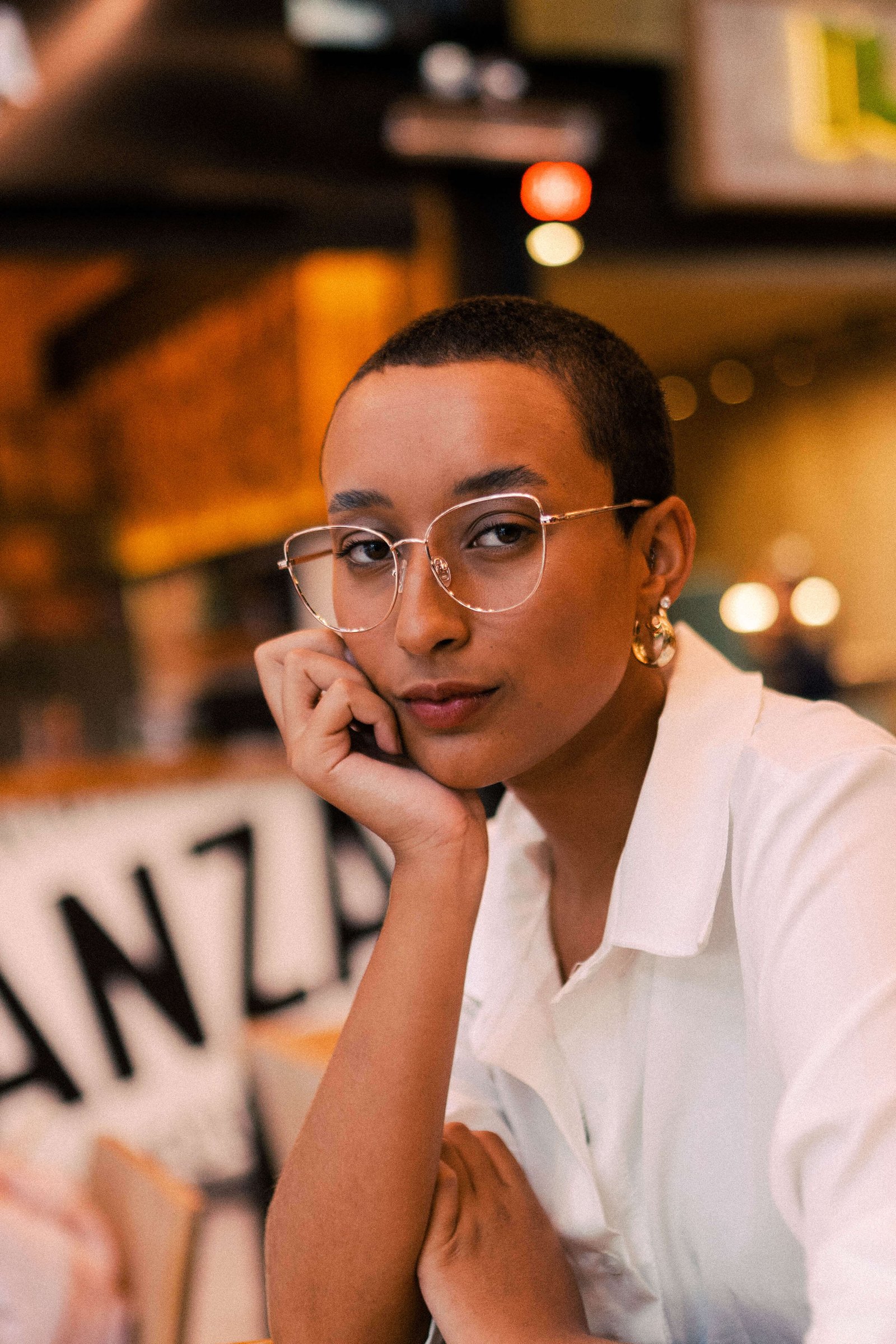 Mulher com cabelo curto e óculos de armação transparente apoiando o rosto na mão em cafeteria.