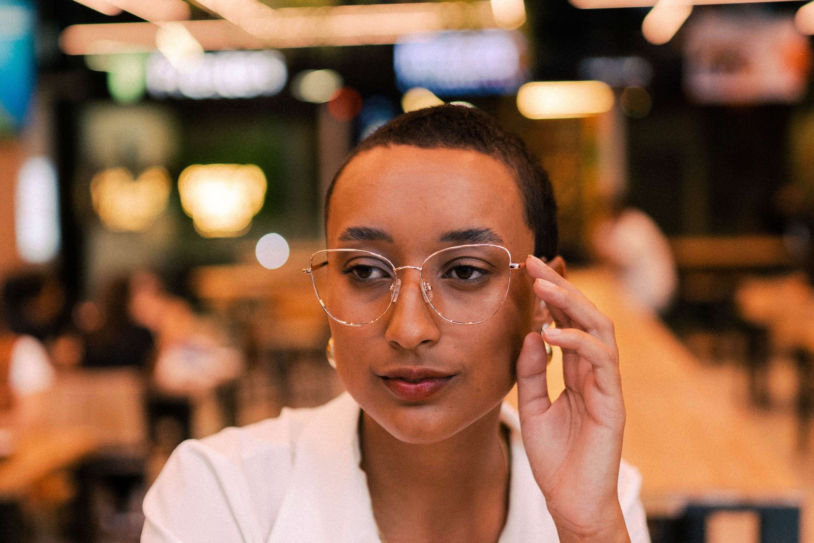 Mulher com óculos de grau olhando pensativa para o lado, sentada em área externa de café.