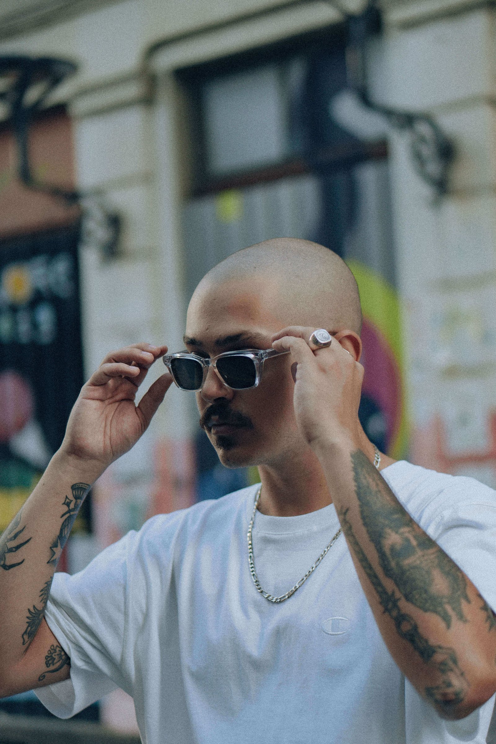 Homem de perfil colocando óculos de sol, com fachada de prédio e cartazes desfocados ao fundo.