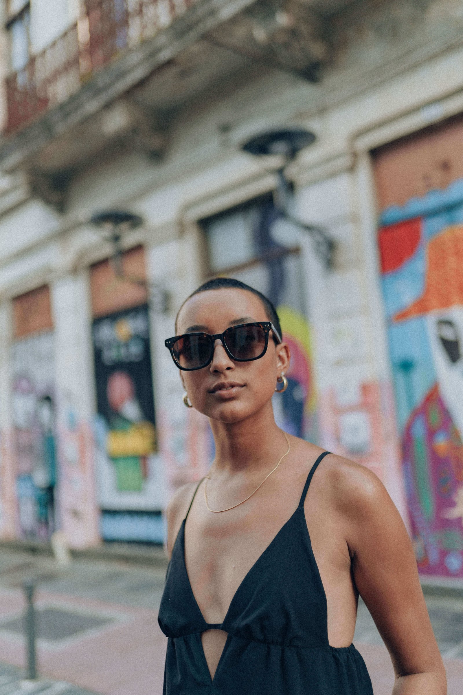 Mulher de vestido preto e óculos de sol grandes caminhando em calçada com fachadas coloridas ao fundo.