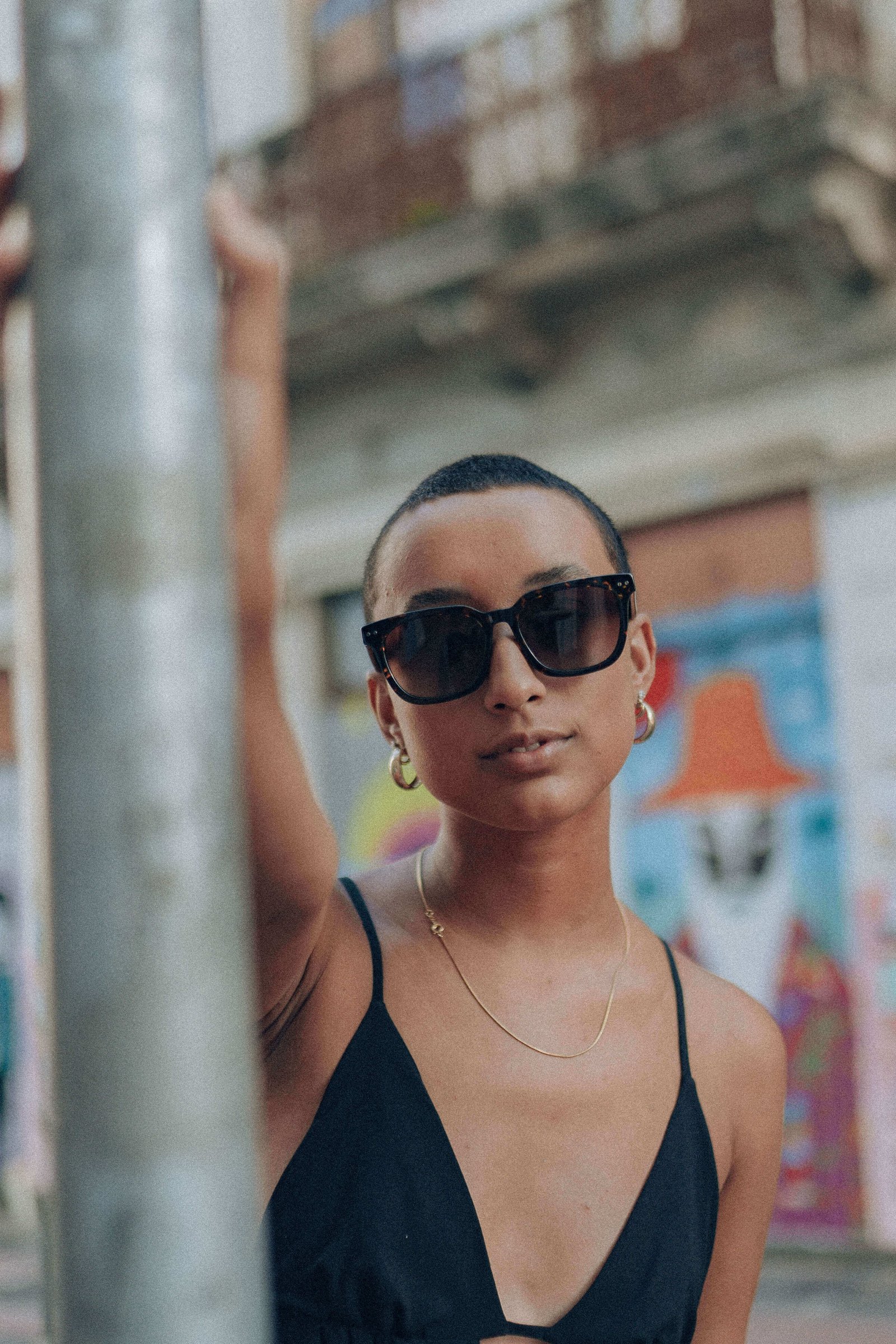 Close da mulher de vestido preto encostada em um poste, usando óculos de sol pretos e expressão confiante.