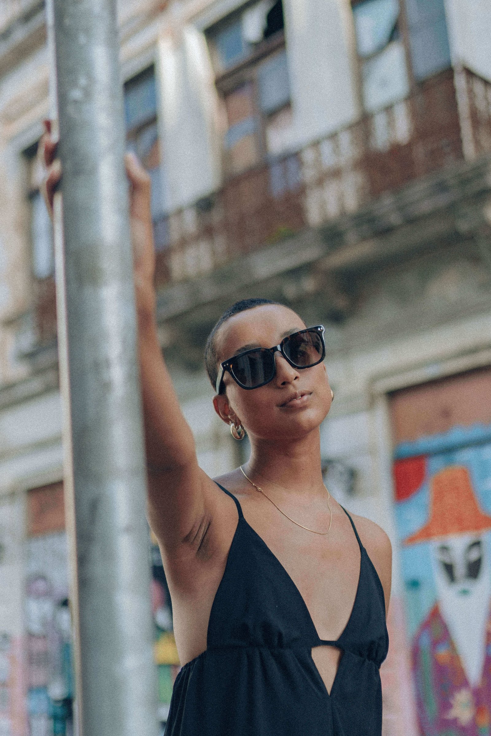 Mulher com vestido preto e óculos de sol arredondados levantando o braço ao lado de um poste em rua urbana.