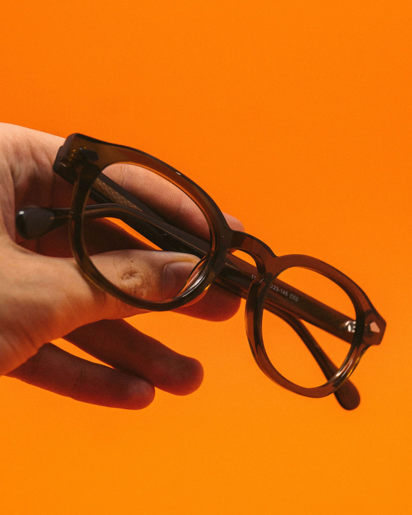 Mão segurando óculos de grau preto em frente a fundo laranja vibrante.