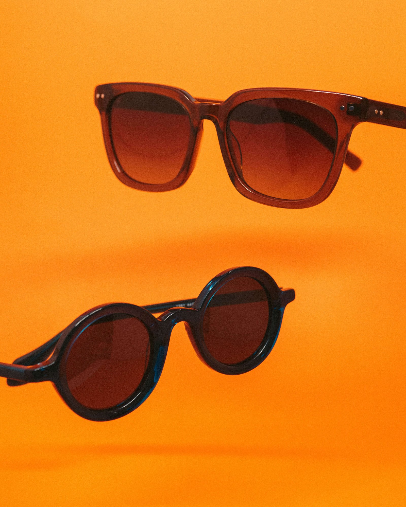 Dois óculos de sol escuros em fundo laranja, um visto de frente e outro de lado.